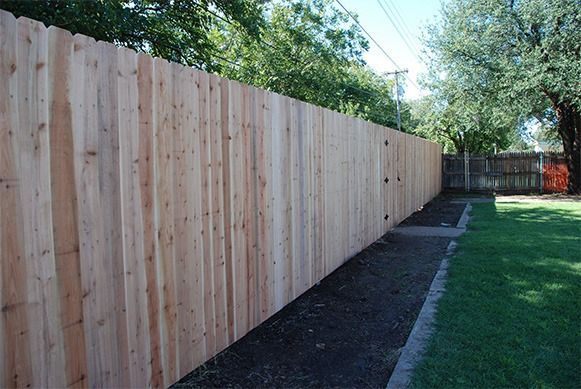 Tall wooden privacy fence along a grassy yard with a narrow dirt border and trees overhead