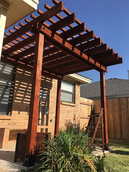 Wooden pergola beside a brick house, with a ladder leaning against it in a backyard.
