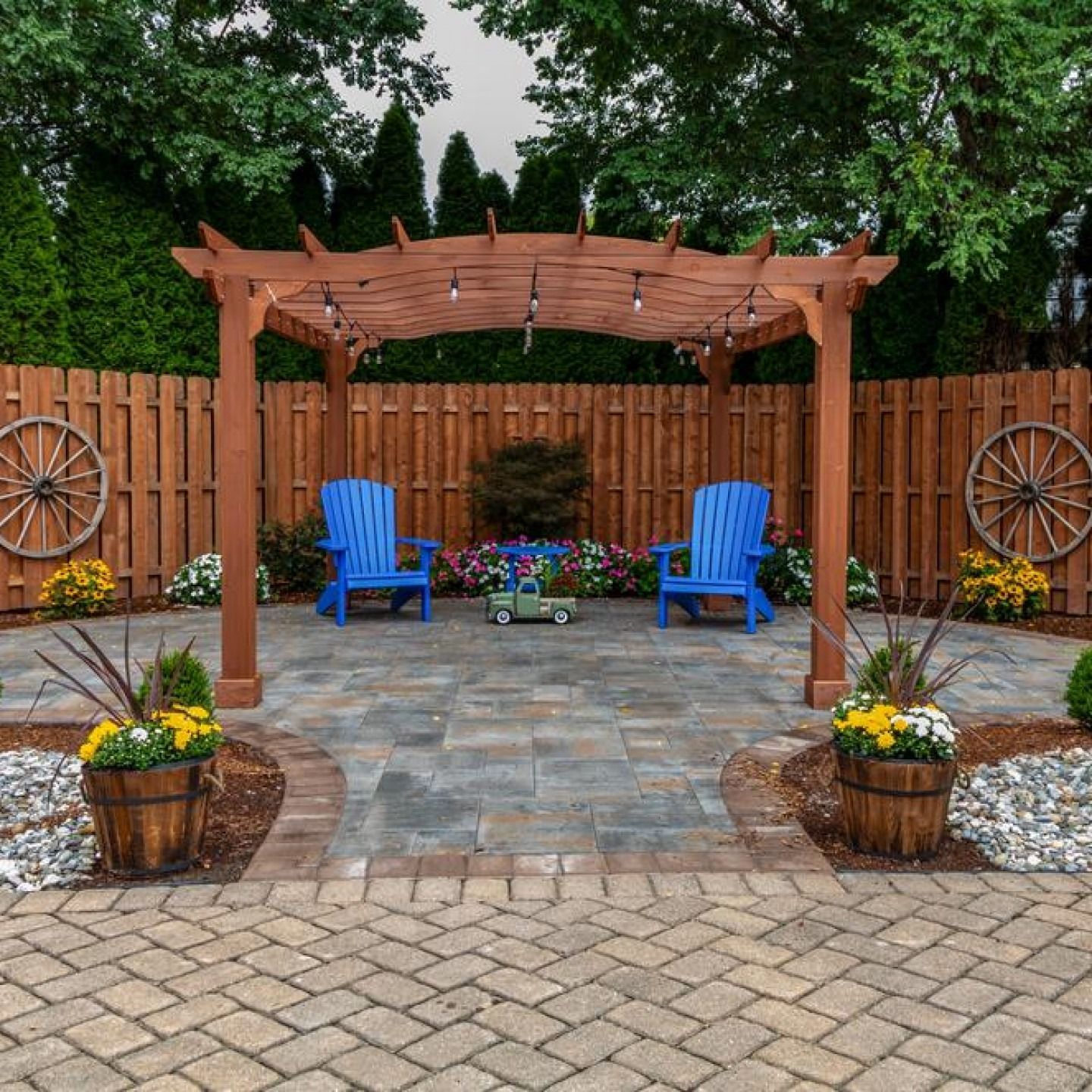 Backyard patio with wooden pergola, two blue chairs, flower pots, and stone paving beneath trees