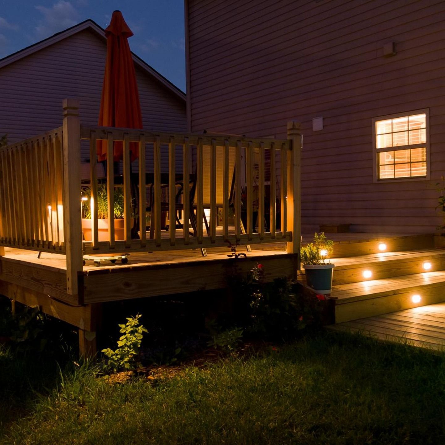 Backyard deck lit at night with wooden railing, orange umbrella, and steps glowing by small lights
