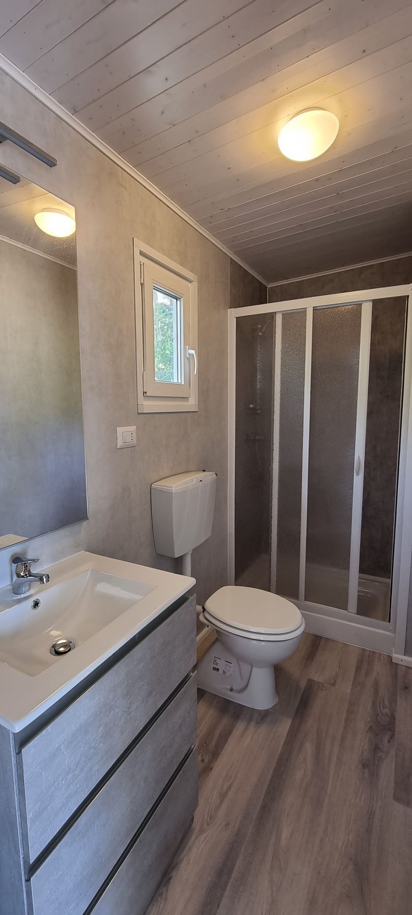 Badkamer met wastafel, toilet en douche. Grijze muren, vloer met houtlook en een raam.