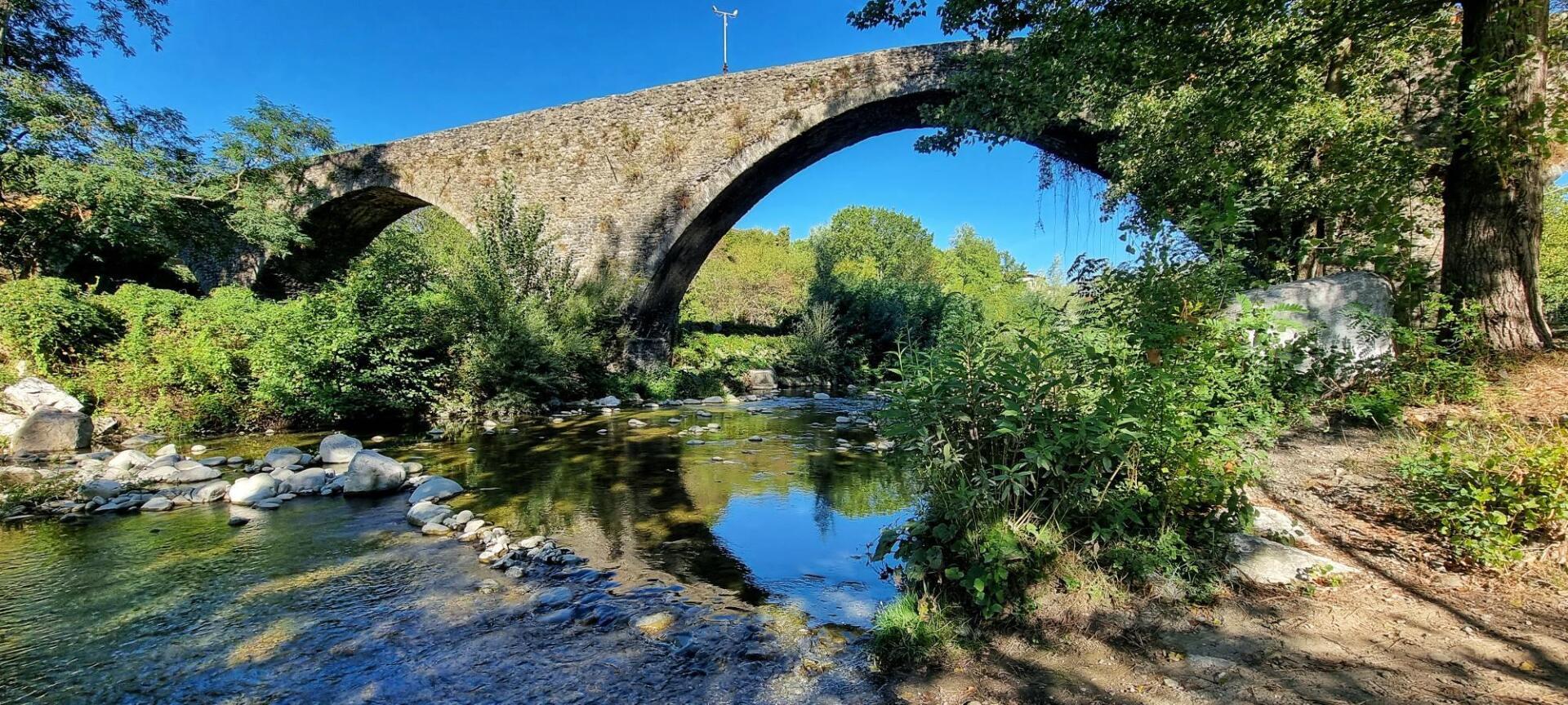 Een stenen boogbrug over een ondiepe rivier, met een heldere hemel en weelderige groene vegetatie. Pontremoli