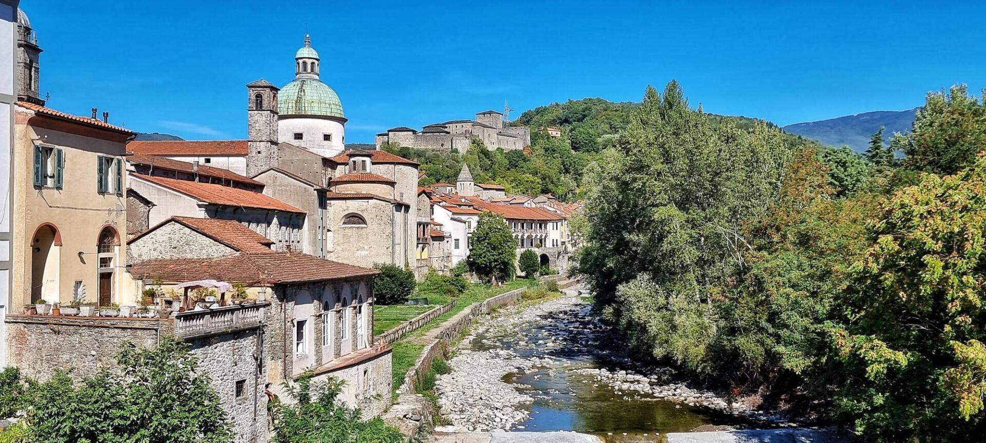 Pontremoli met stenen gebouwen, rode pannendaken, een rivier en een kasteel op een heuveltop, onder een helderblauwe hemel.
