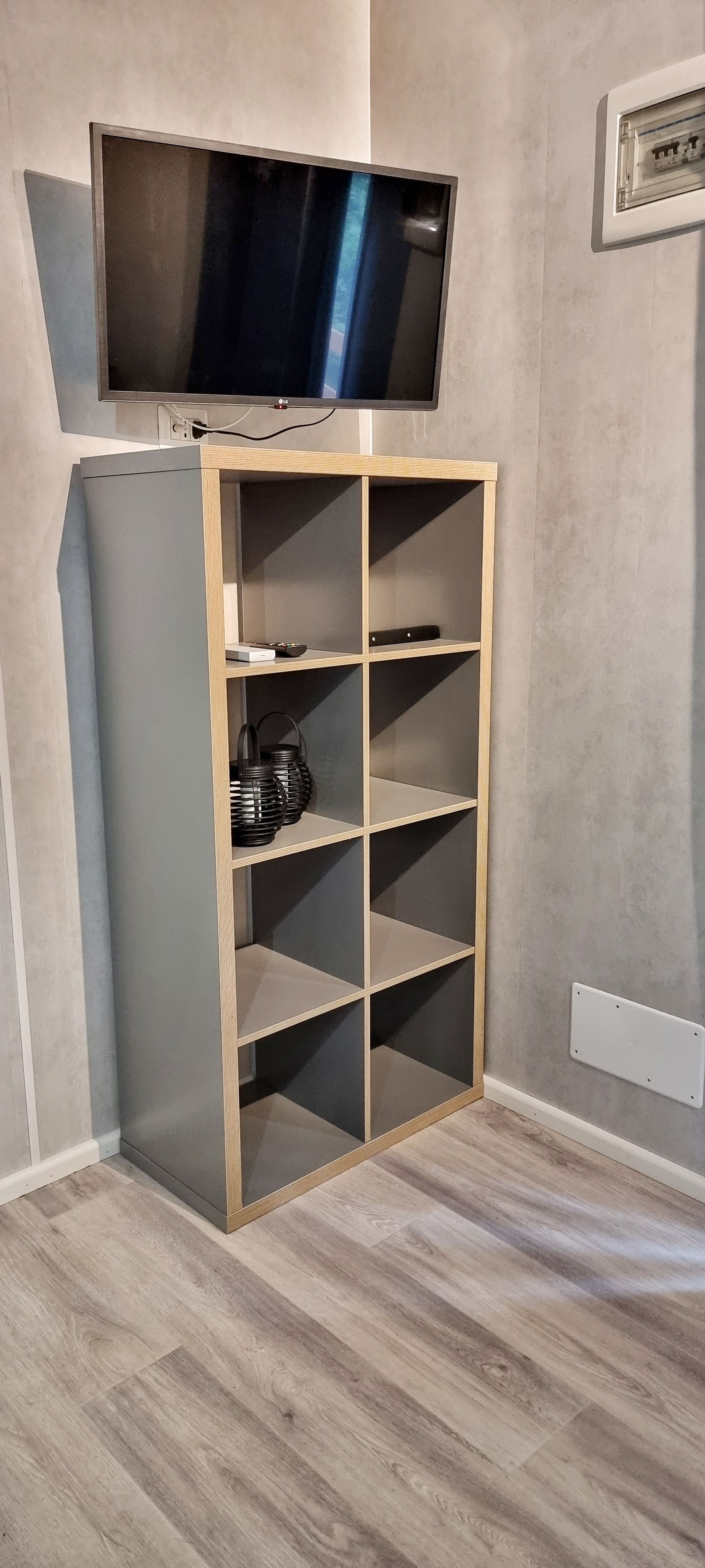Een tv is gemonteerd boven een grijs kubusvormig schap in de hoek van een kamer met een vloer in houtlook.