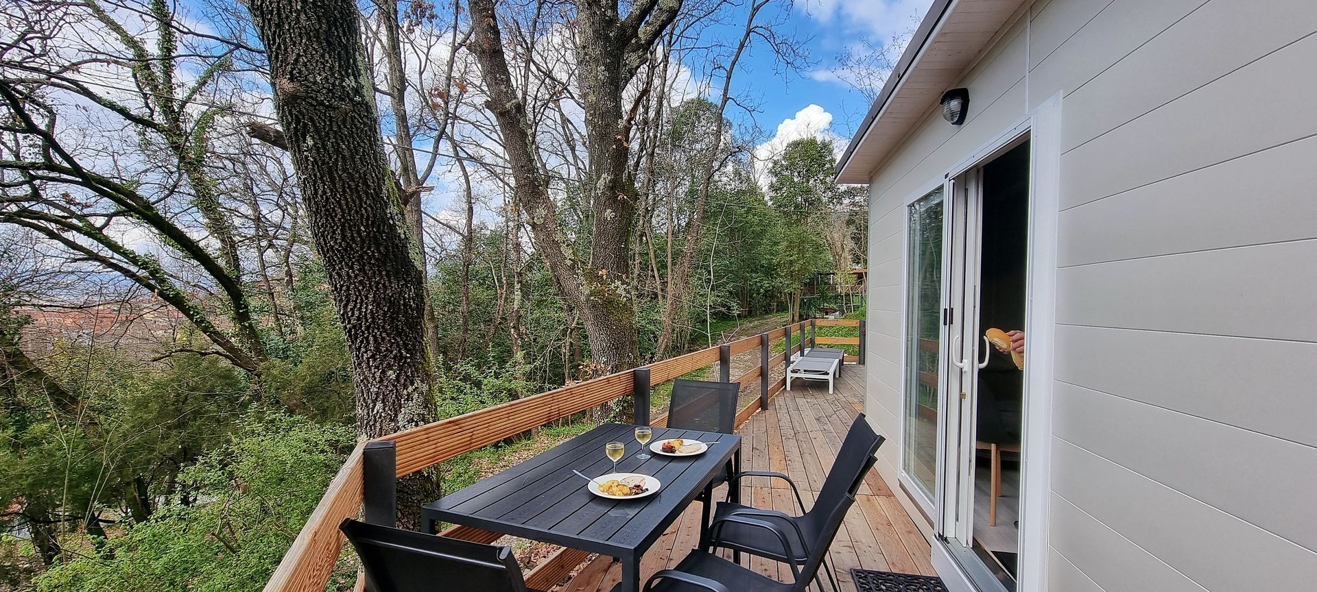 Een houten terras met een tafel en stoelen, met uitzicht op een bos. Het huis heeft schuifdeuren van glas.