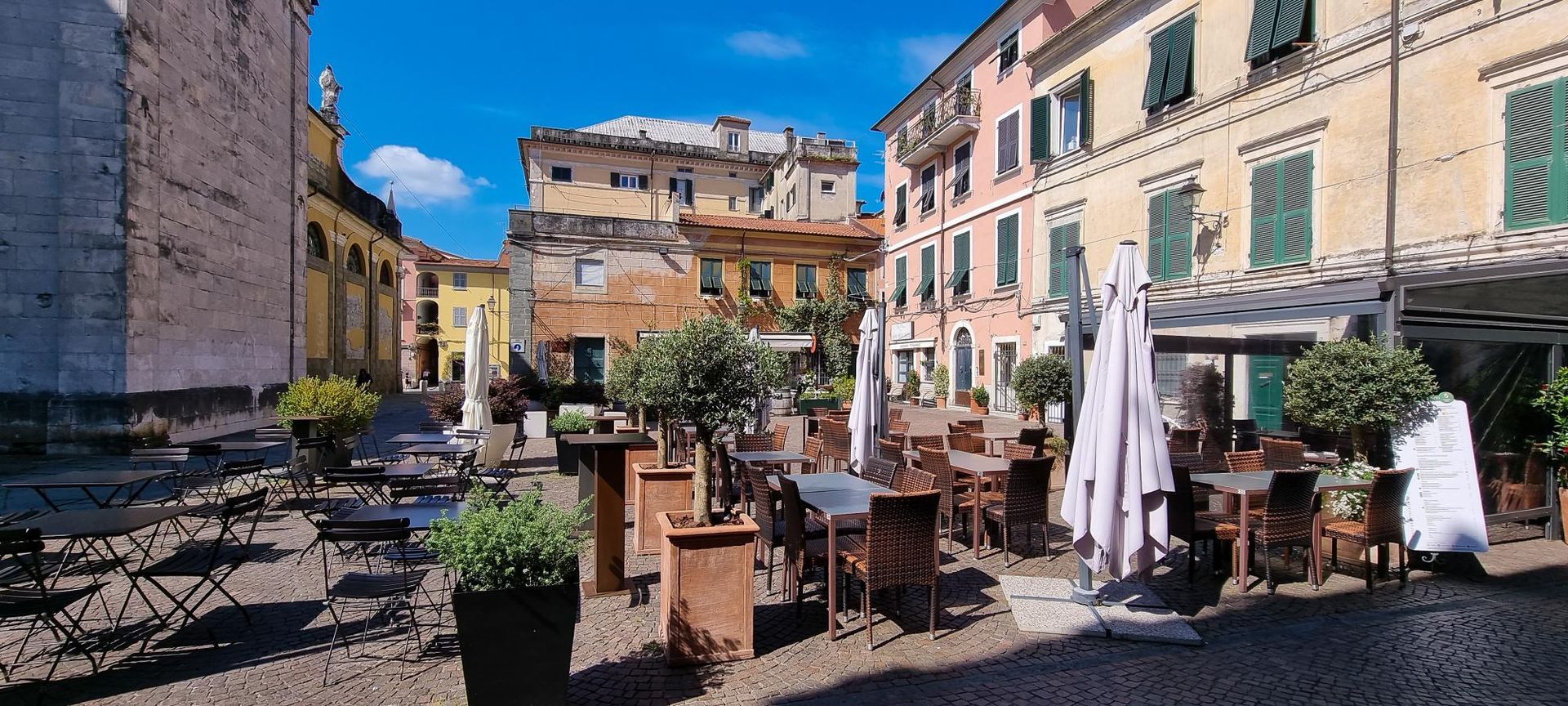 Een terrasje met tafels en parasols op een Europees stadsplein met kleurrijke gebouwen en een blauwe hemel. sarzana