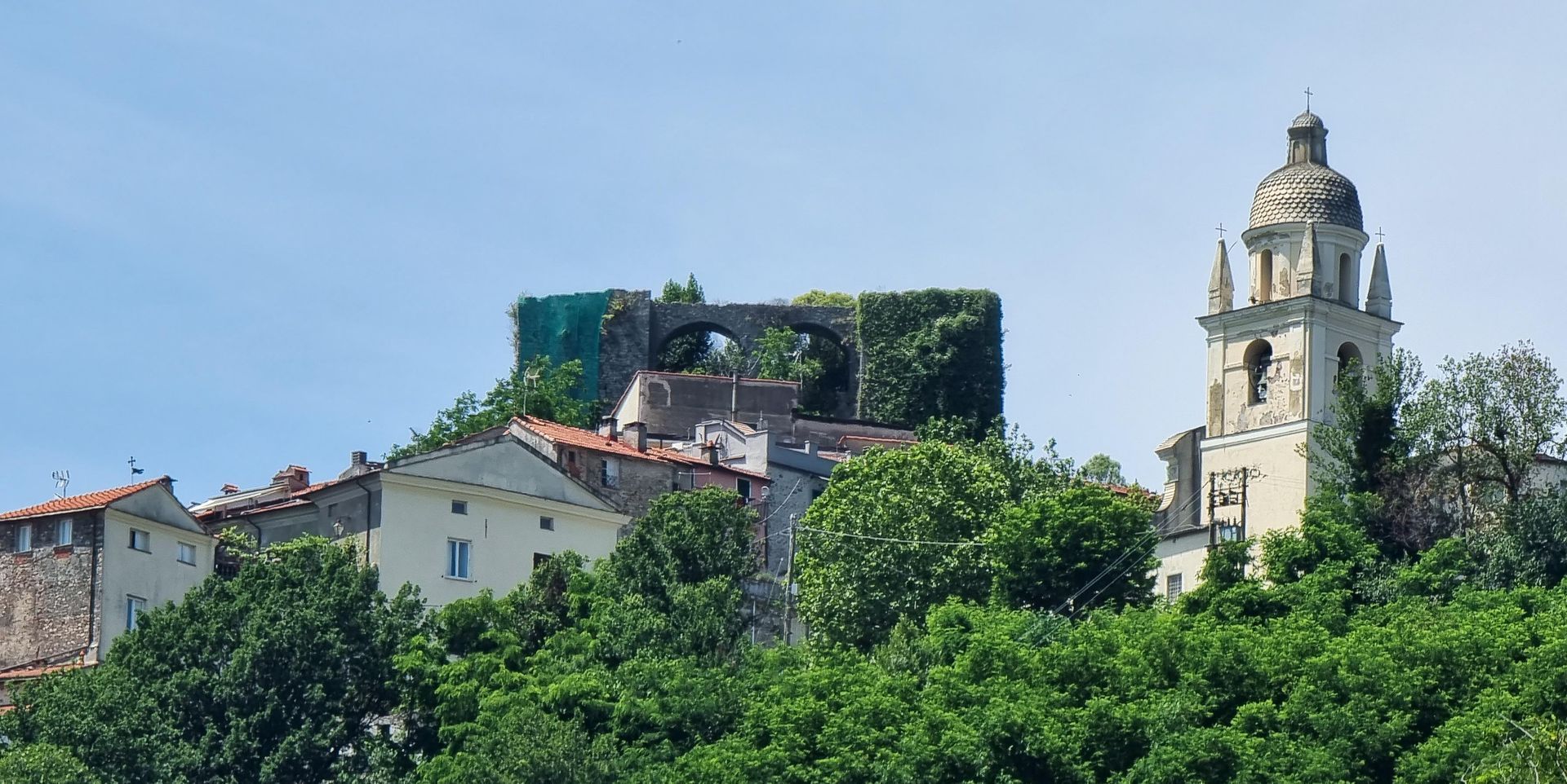 Een dorpje op een heuveltop met stenen gebouwen en een hoge kerktoren tegen een blauwe hemel. Trebiano