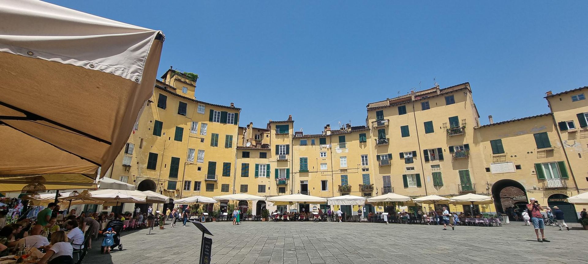 Plein met gele gebouwen en terrasjes onder een helderblauwe hemel. Mensen zitten en lopen. Lucca