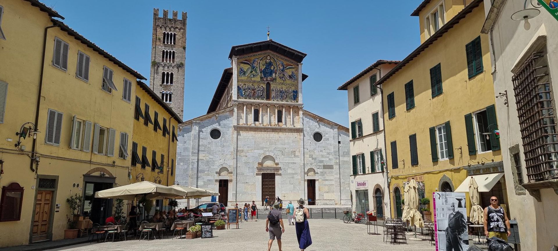 Een plein in Lucca, Italië, met een witte stenen kerk en een hoge klokkentoren.