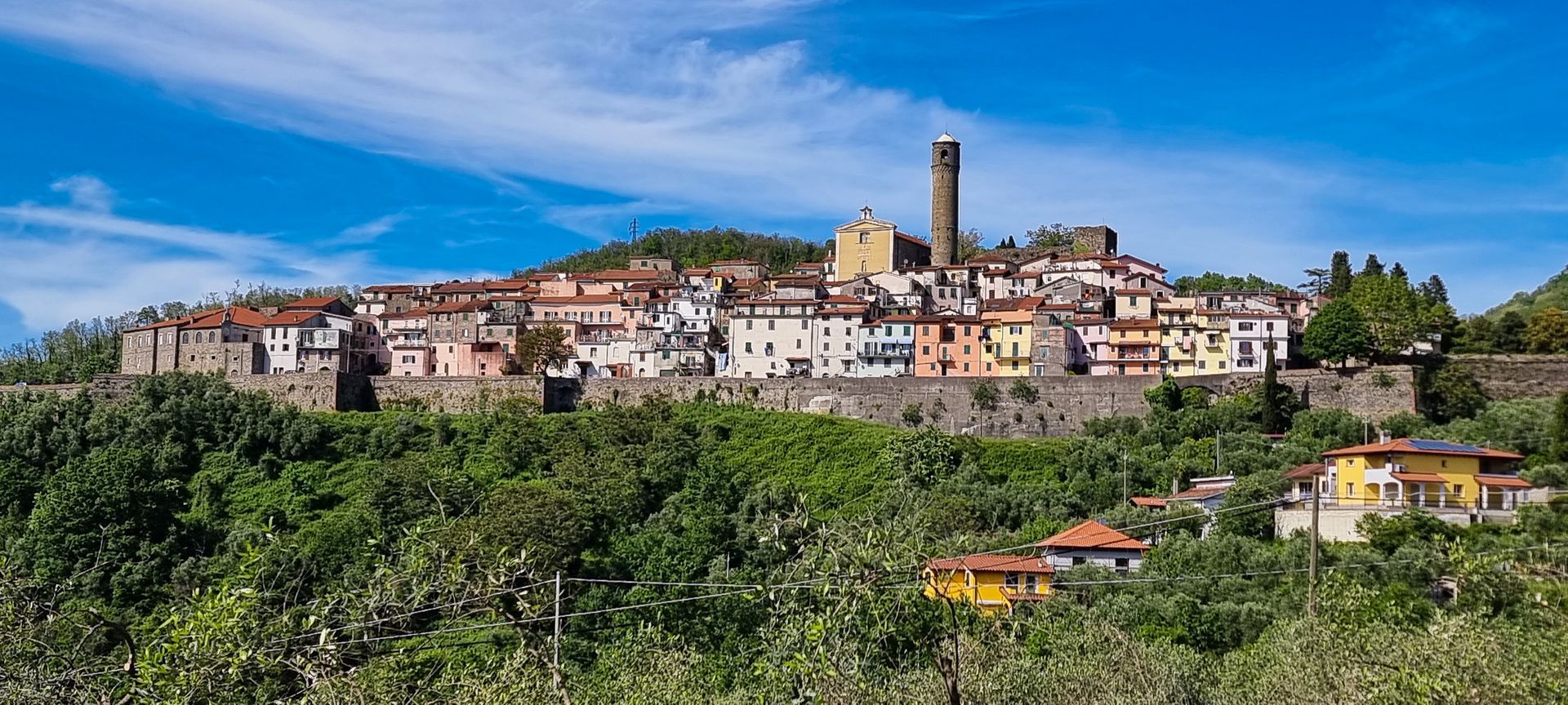 Een kleurrijk Italiaans stadje ligt bovenop een heuvel, omgeven door groene bomen onder een helderblauwe hemel.