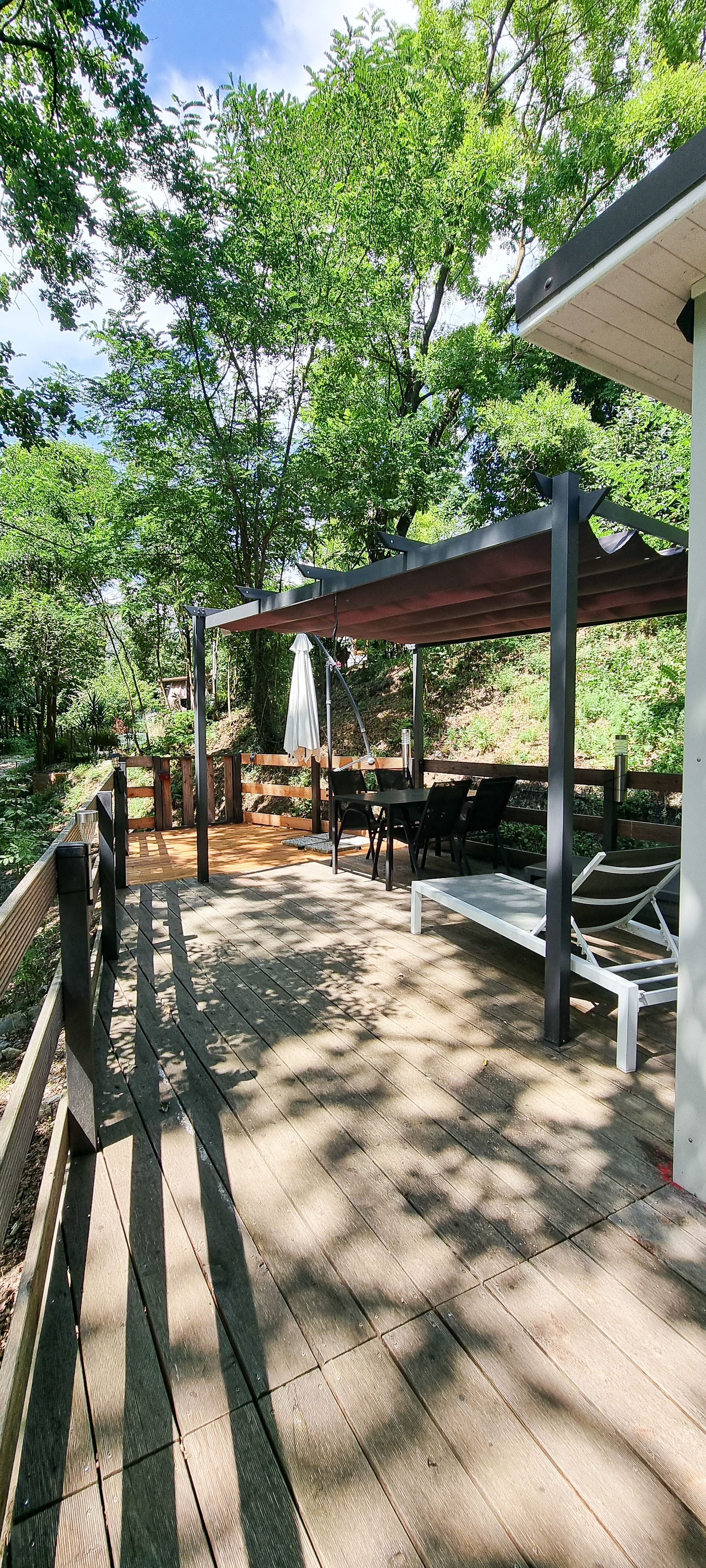Houten terras met pergola, ligstoel en weelderig groen. Zonnige dag, schaduwen op het terras.