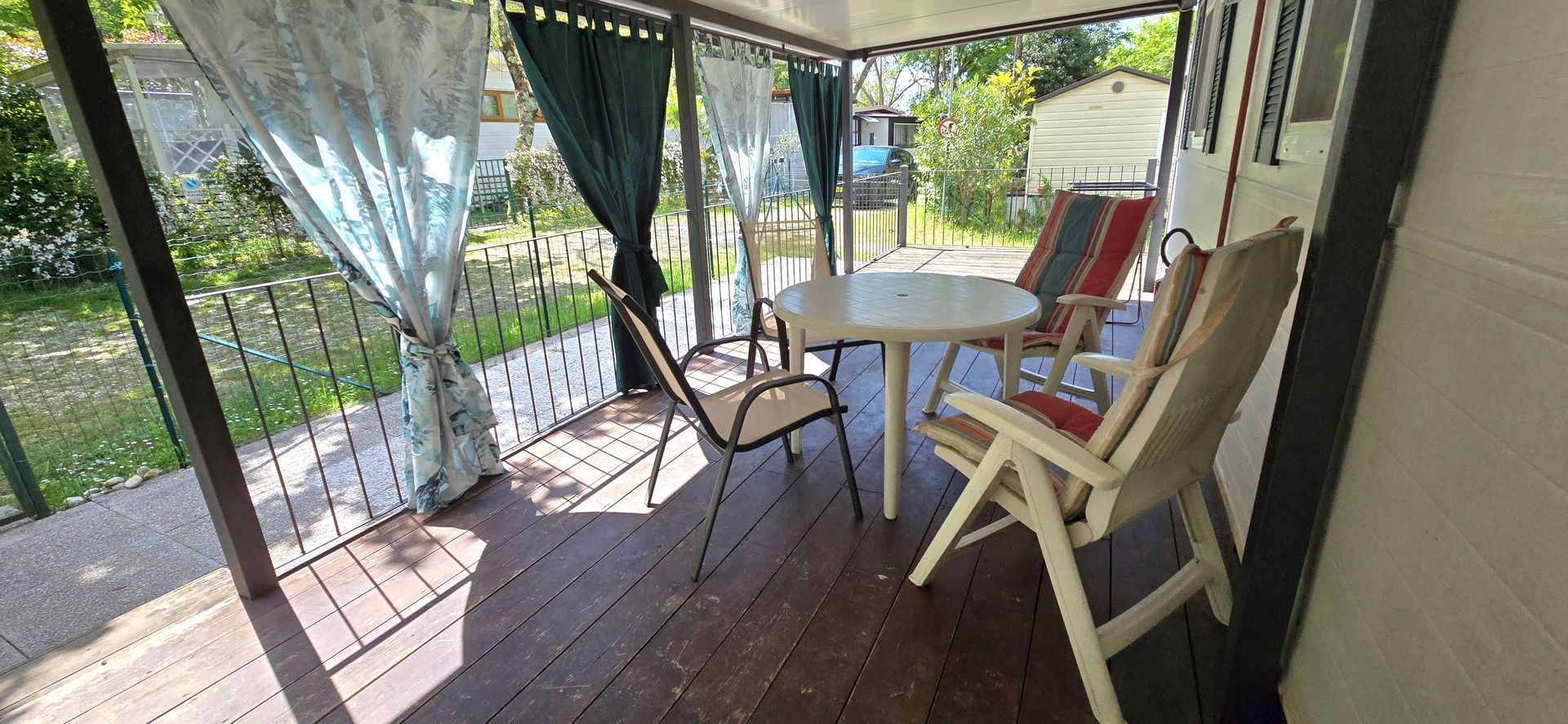 Overdekte veranda met stoelen, tafel en gordijnen. Houten vloer, planten buiten. Zonnige dag.