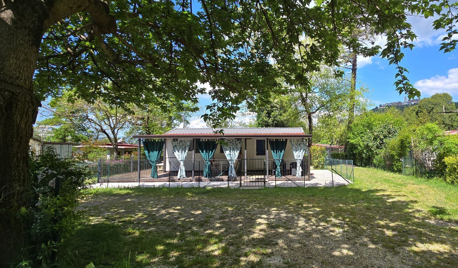 Chalet Anna met turquoise gordijnen op de veranda, omgeven door bomen en een groen gazon, op een zonnige dag.