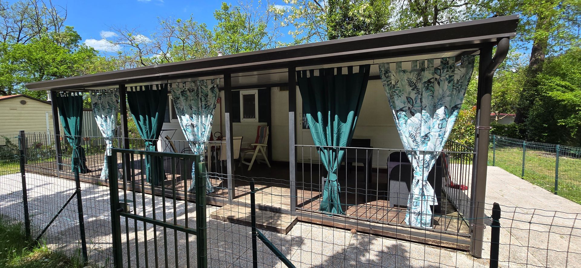 Overdekt terras met gordijnen, zitgelegenheid en een poort. Buiten, vlakbij bomen.