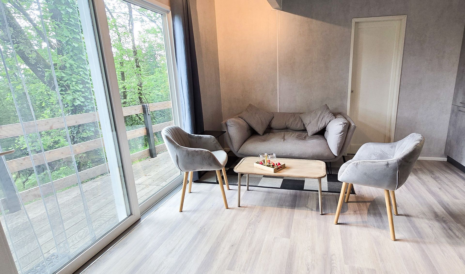 Woonkamer met een grijze bank, twee stoelen en een klein tafeltje bij grote ramen met uitzicht op bomen.