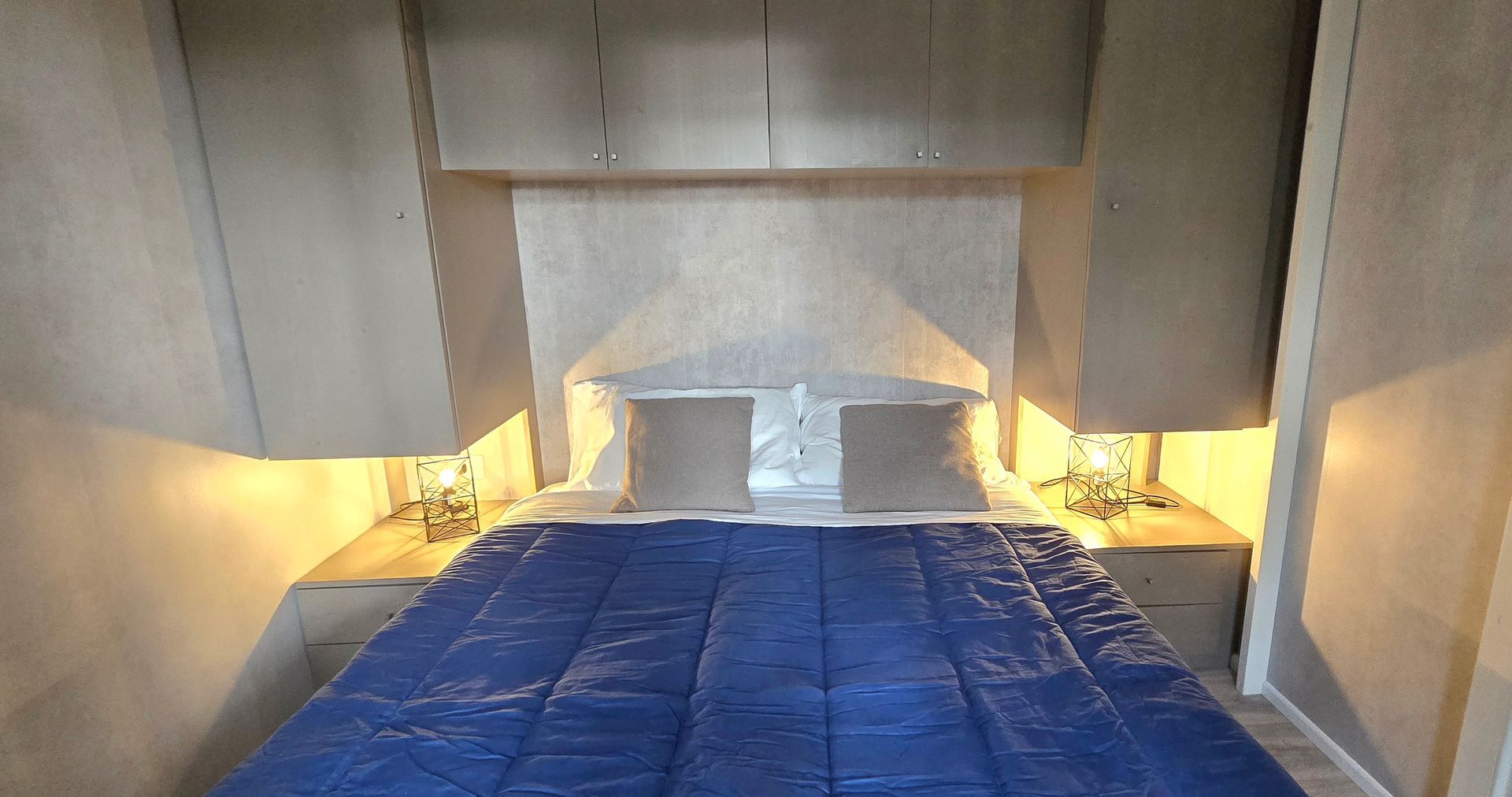 Slaapkamer met 2 persoons bed , nachtkastjes en opbergruimte boven het bed; blauwe deken, grijze muren en zachte verlichting.