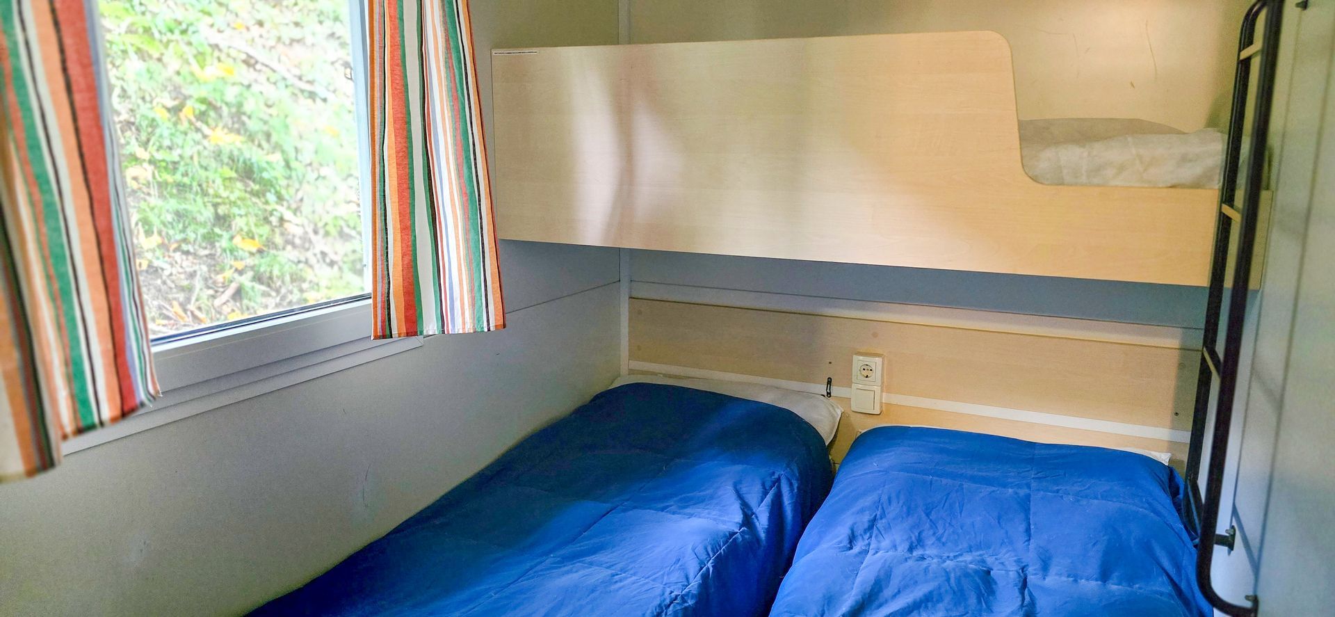 Stapelbedden in een kleine kamer met blauw beddengoed, een raam met gestreepte gordijnen en een ladder.