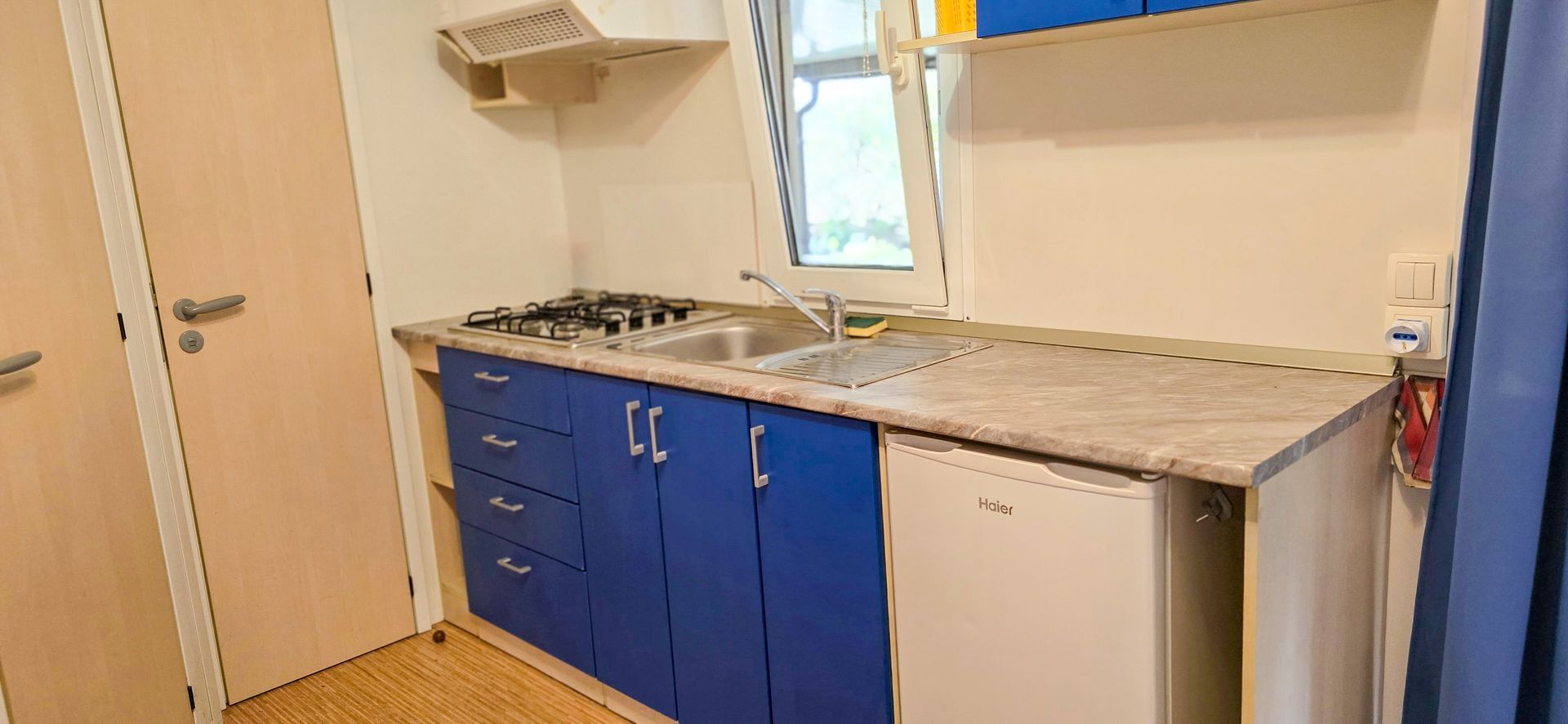 Keuken met blauwe kastjes, gasfornuis, spoelbak en koelkast. Lichte houten deuren aan de linkerkant, raam op de achtergrond.