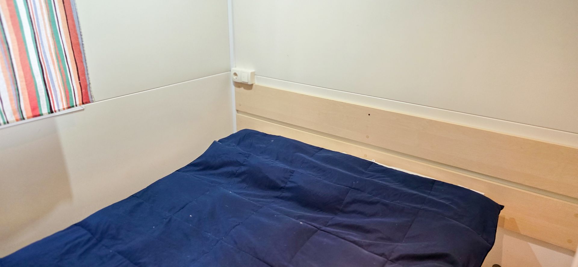 Een kleine, eenvoudige slaapkamer met een blauw dekbed op het bed, crèmekleurige muren en gestreepte gordijnen.