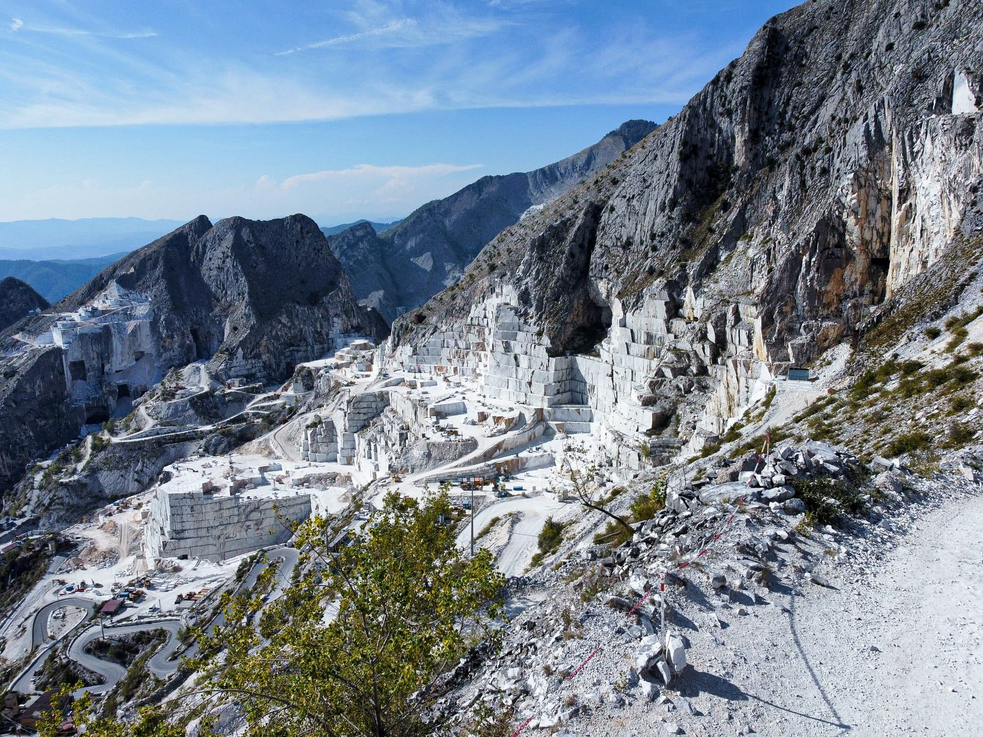 Een marmergroeve uitgehouwen in een berg, met witte rotswanden tegen een blauwe hemel. Carrara