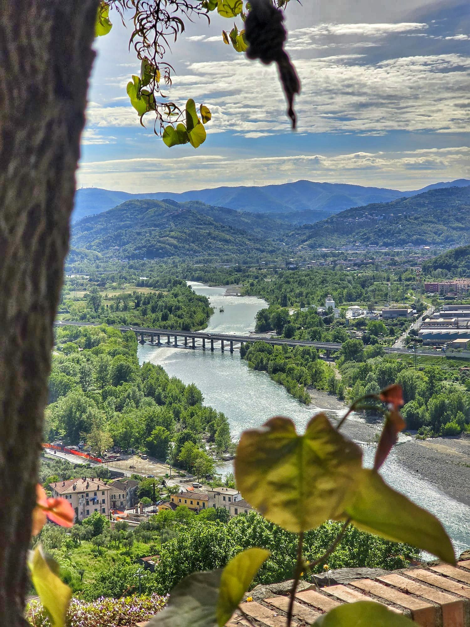 Uitzicht op een vallei met een rivier, een brug, groene bomen, bergen en een blauwe lucht, omlijst door bomen en bladeren.