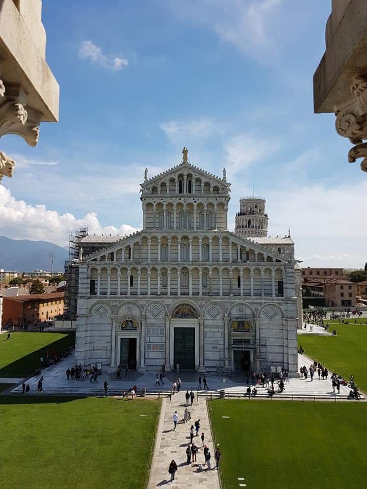 De gevel van de kathedraal van Pisa, wit marmer, mensen lopen over een pad, de scheve toren is zichtbaar, zonnige dag.
