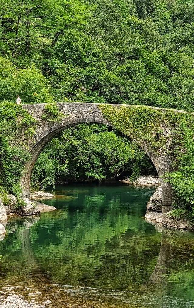 Een stenen boogbrug over een heldere, groene rivier; weelderig groen omringt de rivier.
