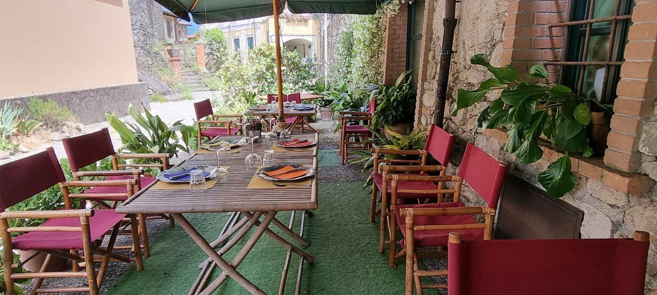 Eetgedeelte buiten met tafels en rode stoelen onder een parasol, omgeven door planten en stenen muren.