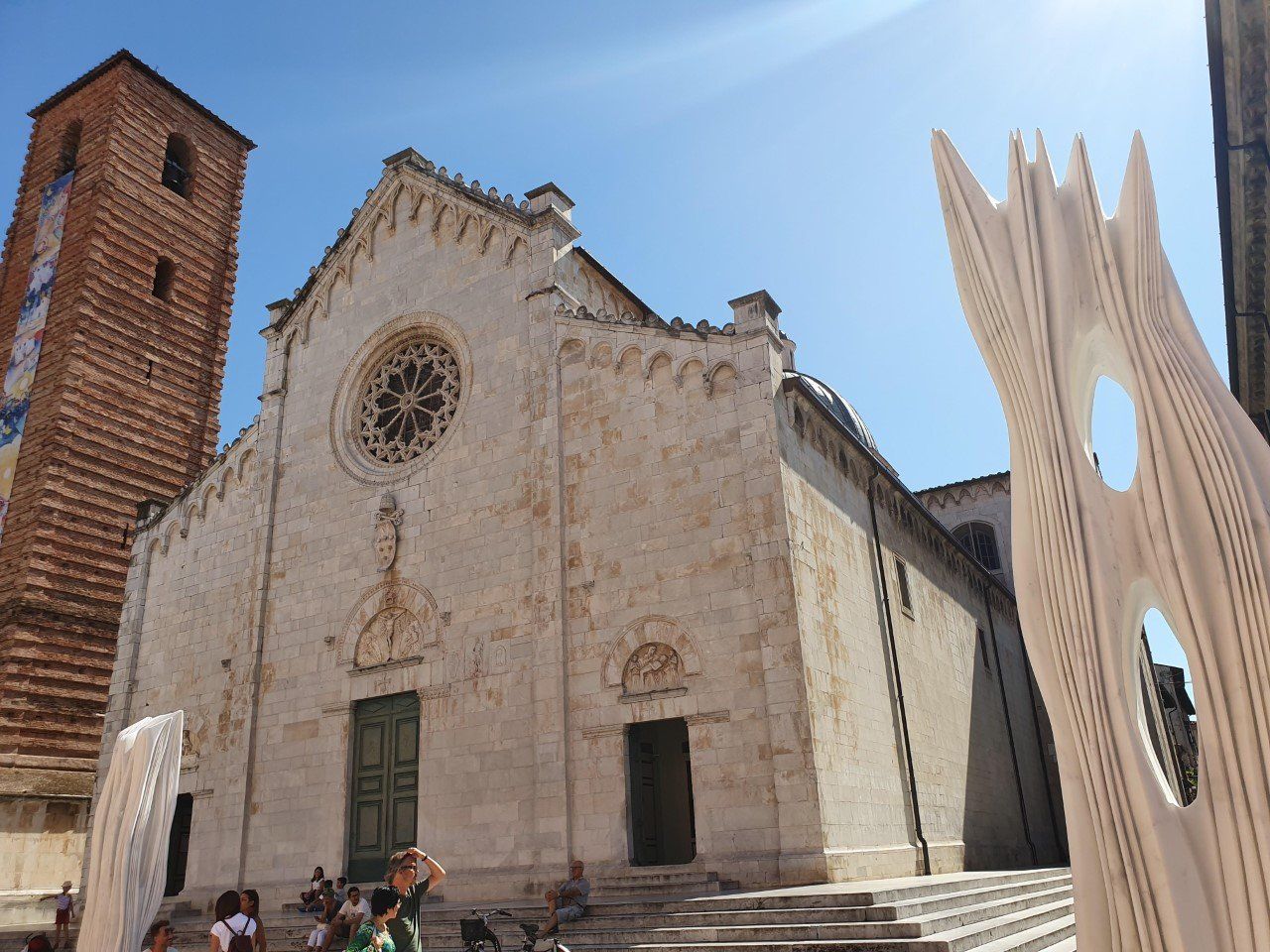 Kerkgevel met roosvenster, klokkentoren en moderne houten sculptuur tegen een blauwe hemel.Pietrasanta