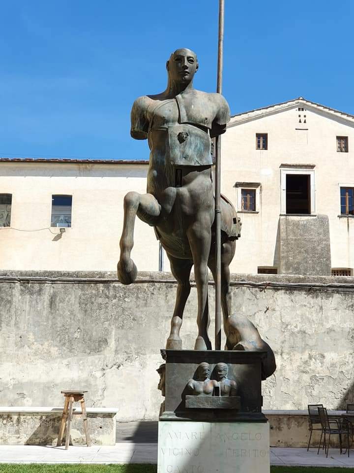 Een centaurbeeld staat buiten een gebouw. Het beeld heeft een bronzen torso. Pietrasanta