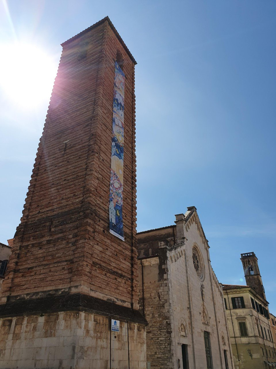 Een hoge bakstenen toren naast een kerk, met de zon die fel aan de hemel schijnt. Pietrasanta