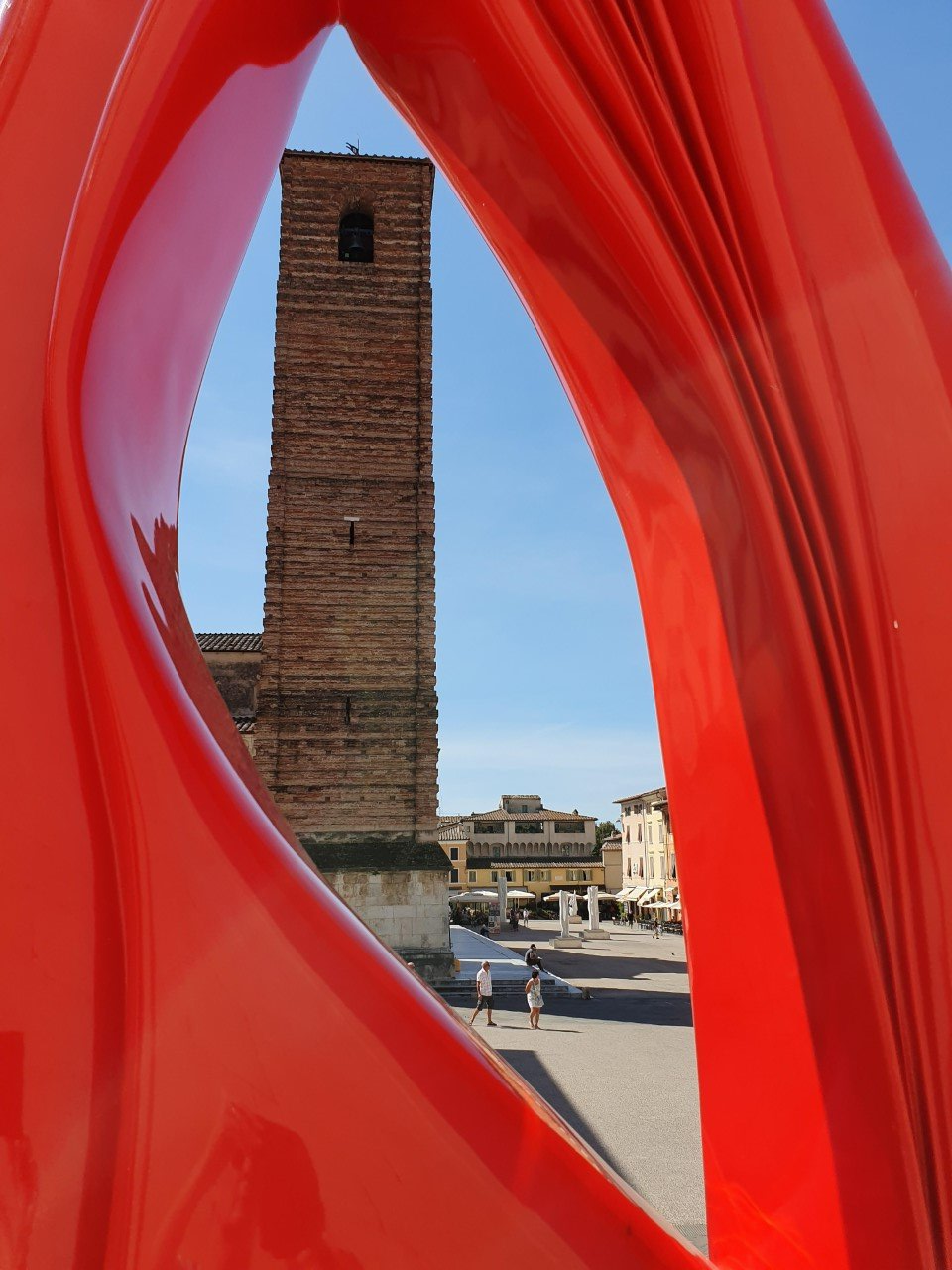 Een bakstenen toren en plein, omlijst door een groot, rood, abstract beeldhouwwerk. Pietrasanta
