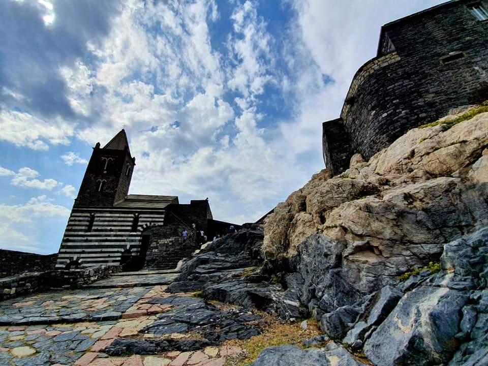Kerk en vesting op een rotsachtige heuvel onder een bewolkte hemel. Zwart-wit gestreepte gevel. Portovenere