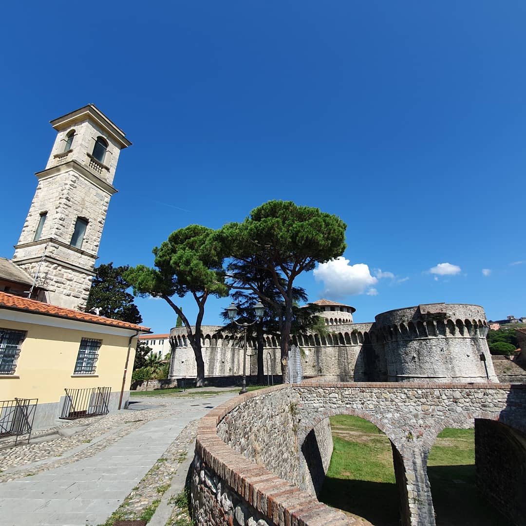 Stenen vesting met een hoge klokkentoren en een boogbrug onder een blauwe hemel. Sarzana