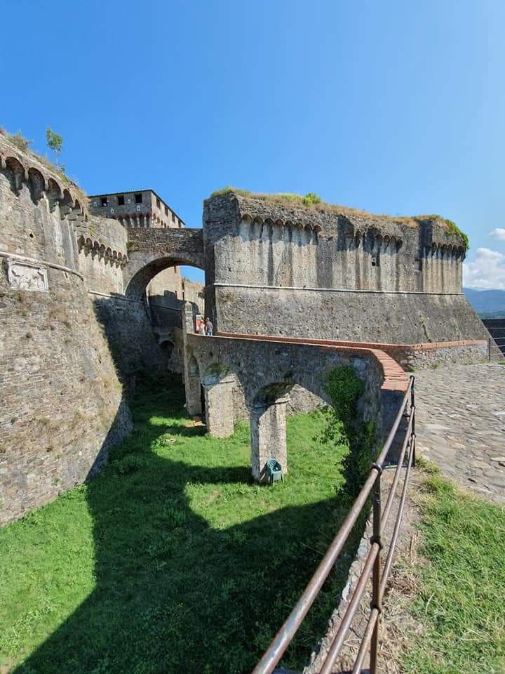 Stenen vesting met bogen, een brug en een groene gracht onder een helderblauwe hemel. Sarzana