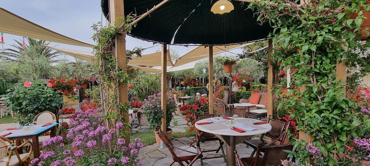 Buitenrestaurant met tafels omgeven door kleurrijke bloemen en groen onder een schaduwrijk afdak.