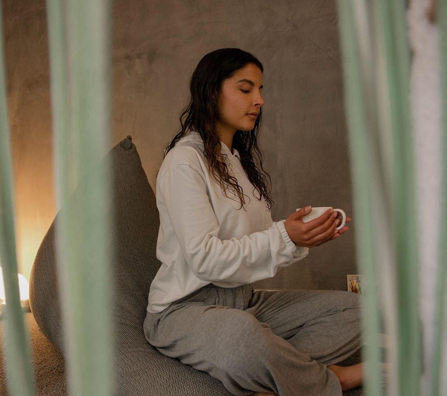 Una mujer con una camiseta blanca y pantalones grises está sentada, sosteniendo una taza, con los ojos cerrados, en un ambiente interior sereno.