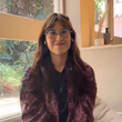 Una mujer con gafas sonríe, sentada junto a una ventana. Lleva una chaqueta morada de lentejuelas y una blusa negra.