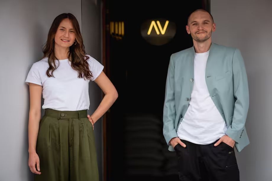 Mujer con camiseta blanca y falda verde, hombre con chaqueta azul, de pie en la puerta con el logo