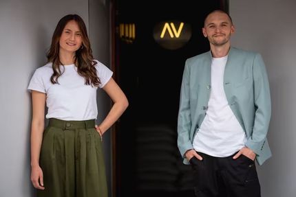 Mujer con camiseta blanca y falda verde, hombre con chaqueta azul, de pie en la puerta con el logo