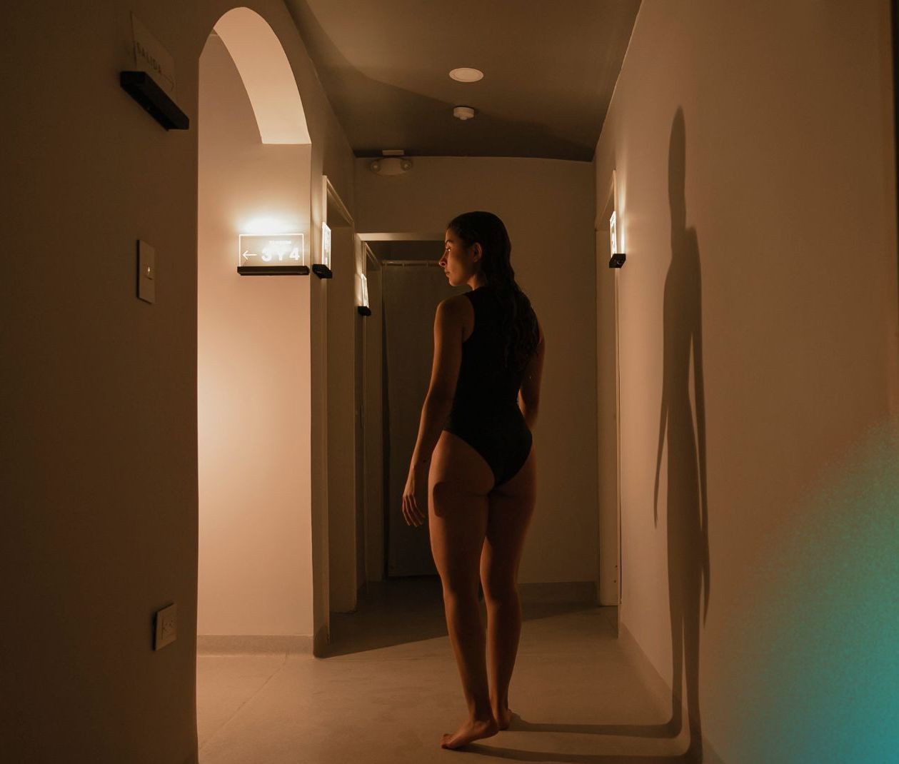 Mujer en traje de baño negro caminando por un pasillo poco iluminado, con sombras en la pared.