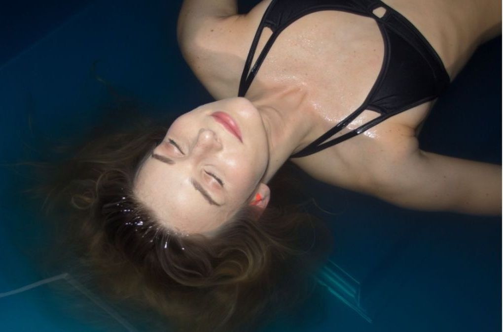 Mujer flotando en agua azul oscuro, con los ojos cerrados y vistiendo un bikini negro.