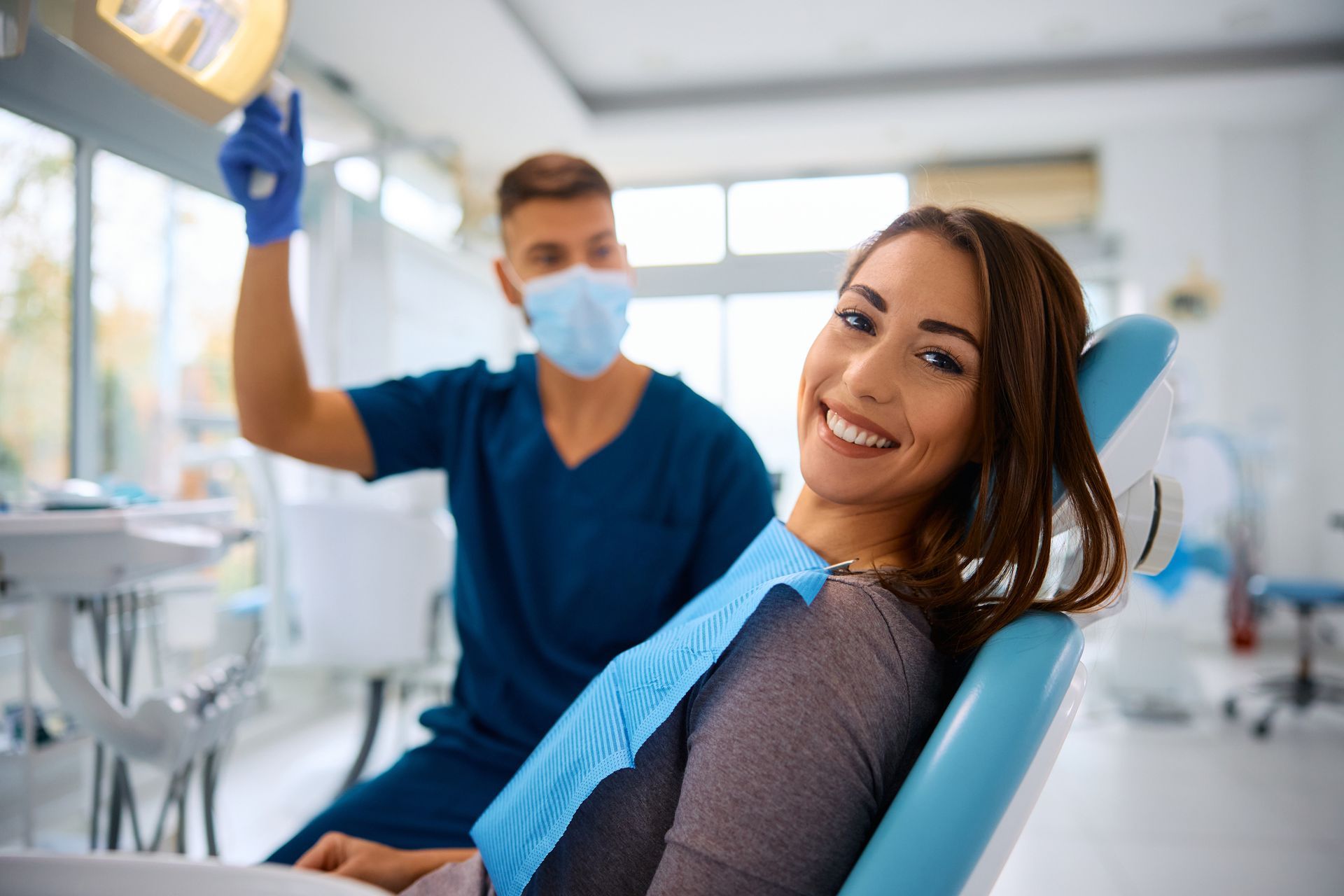 Un dentista esamina i denti di una donna sorridente; si trovano in uno studio medico ed entrambi indossano dispositivi di protezione.
