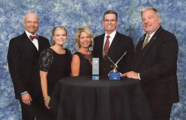 Five people in formal attire posing with awards at a table on a studio backdrop
