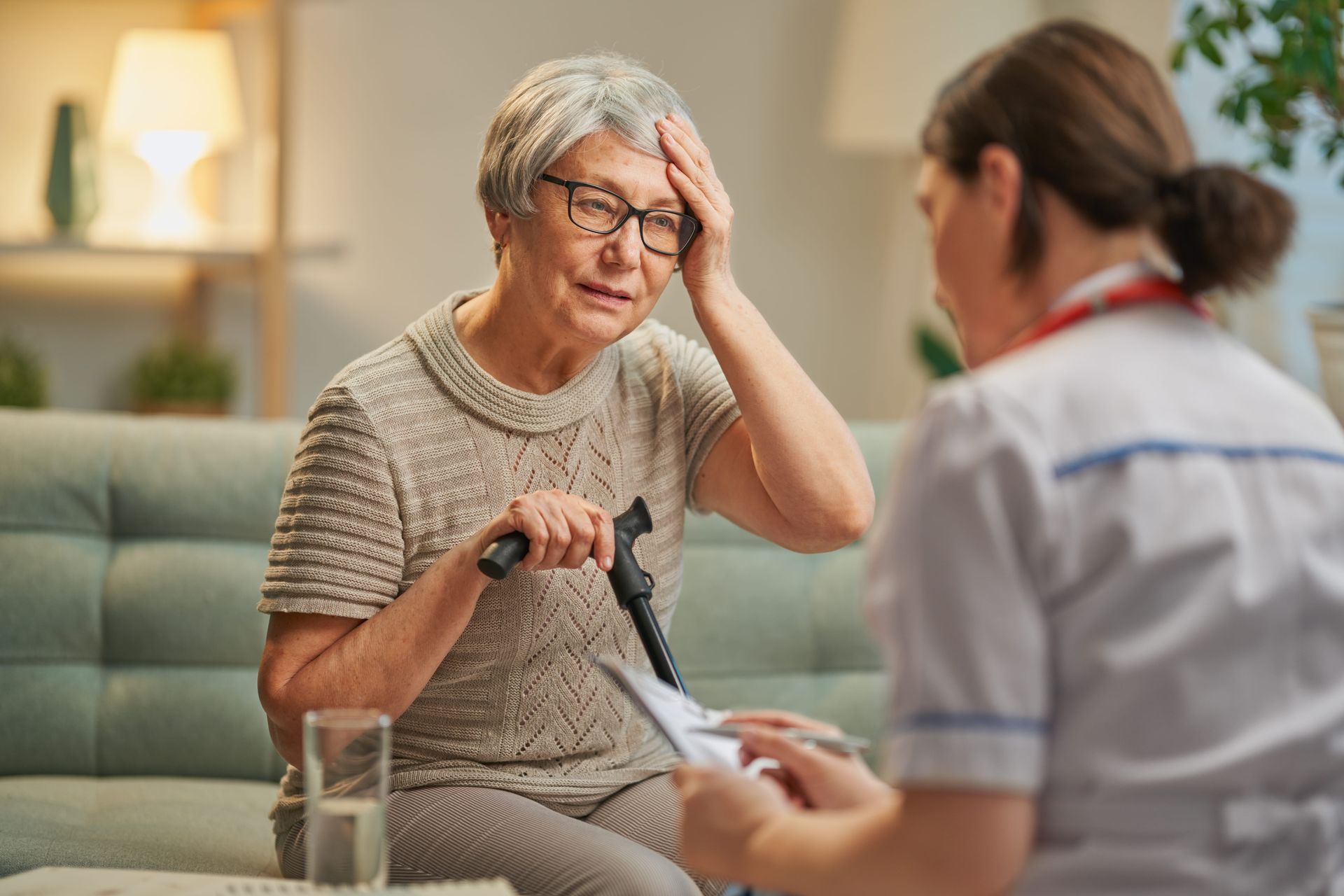 Elderly woman on a couch with a cane, talking with a nurse in a bright living room