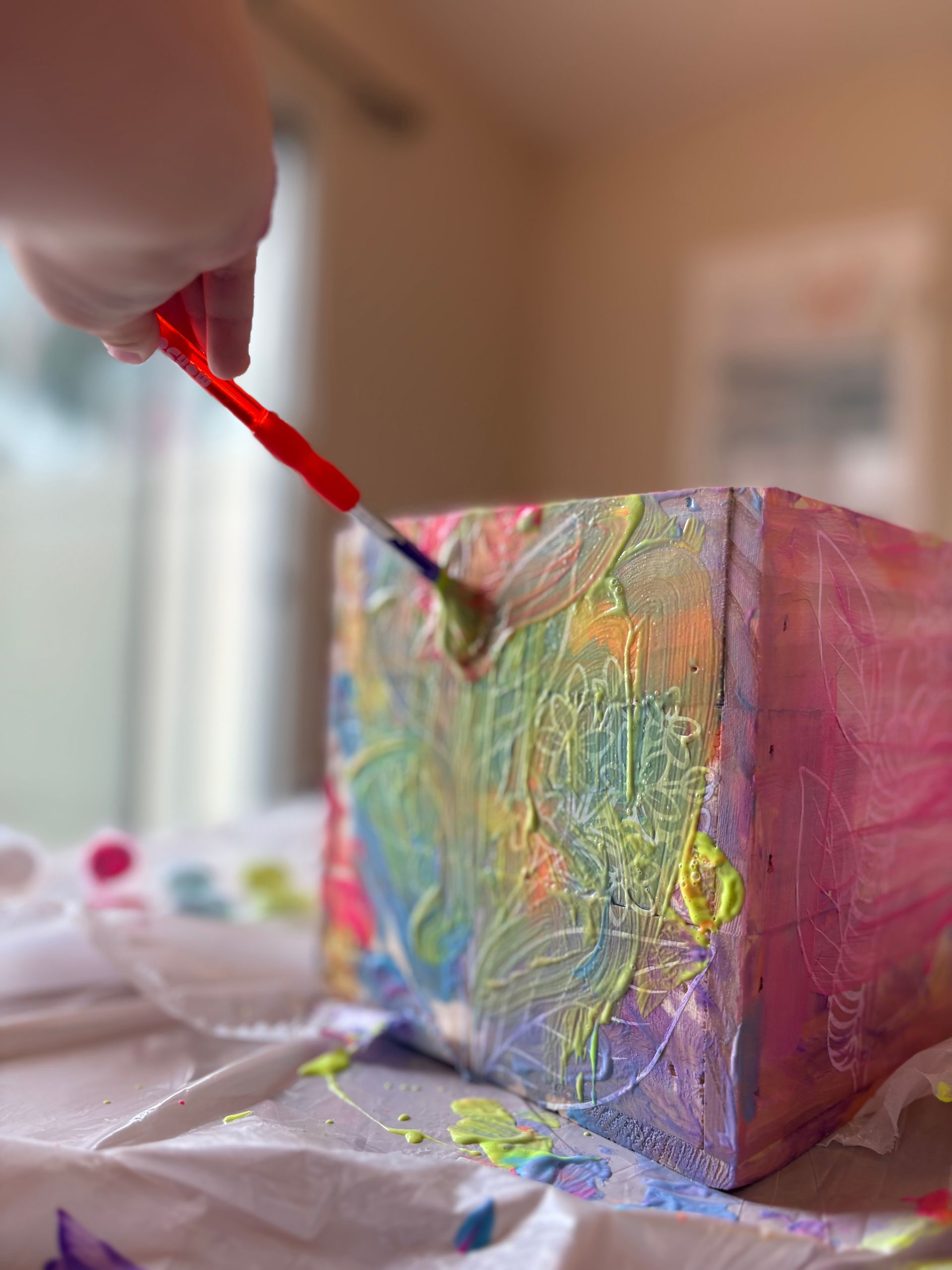 A hand holding a paintbrush applies colorful paint to a textured cube on a white surface.