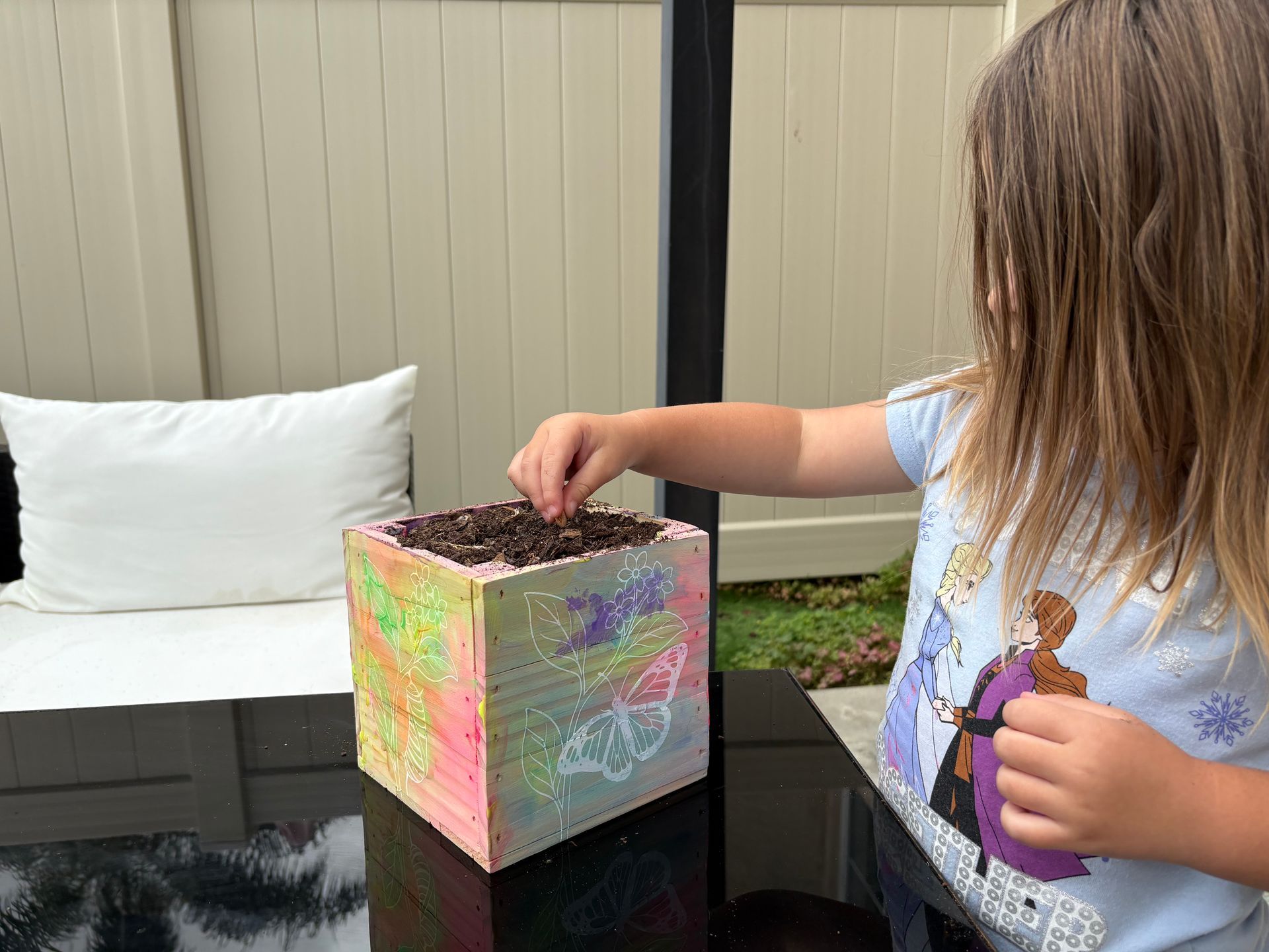 Child planting seeds in a decorated wooden planter on a table outside.