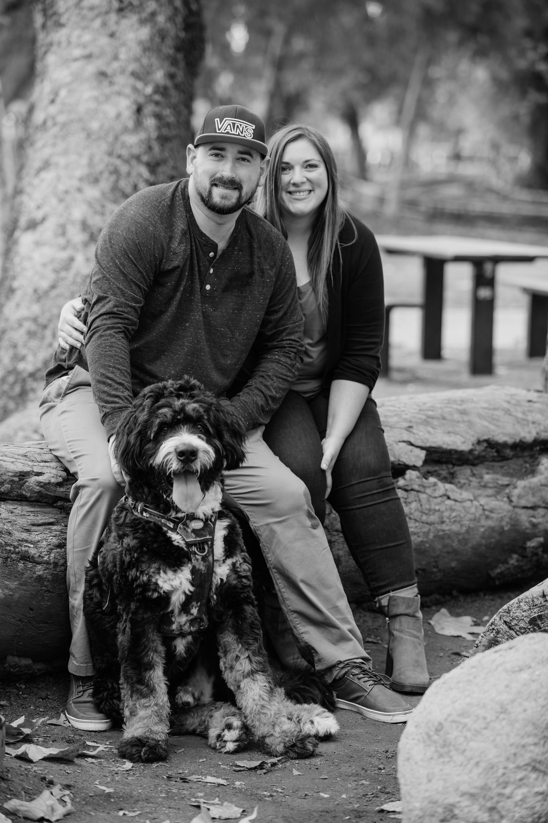 Couple sitting with a large dog outdoors, smiling. Man in a hat, woman in a cardigan.