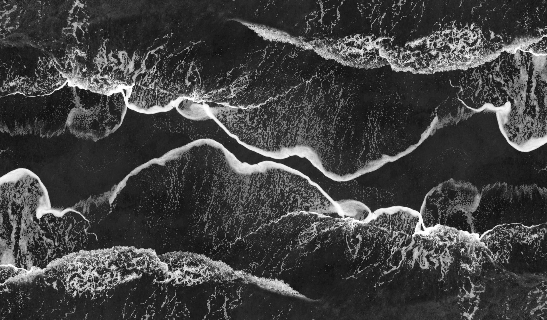Overhead view of ocean waves breaking on a beach, creating foamy white lines against the dark water.