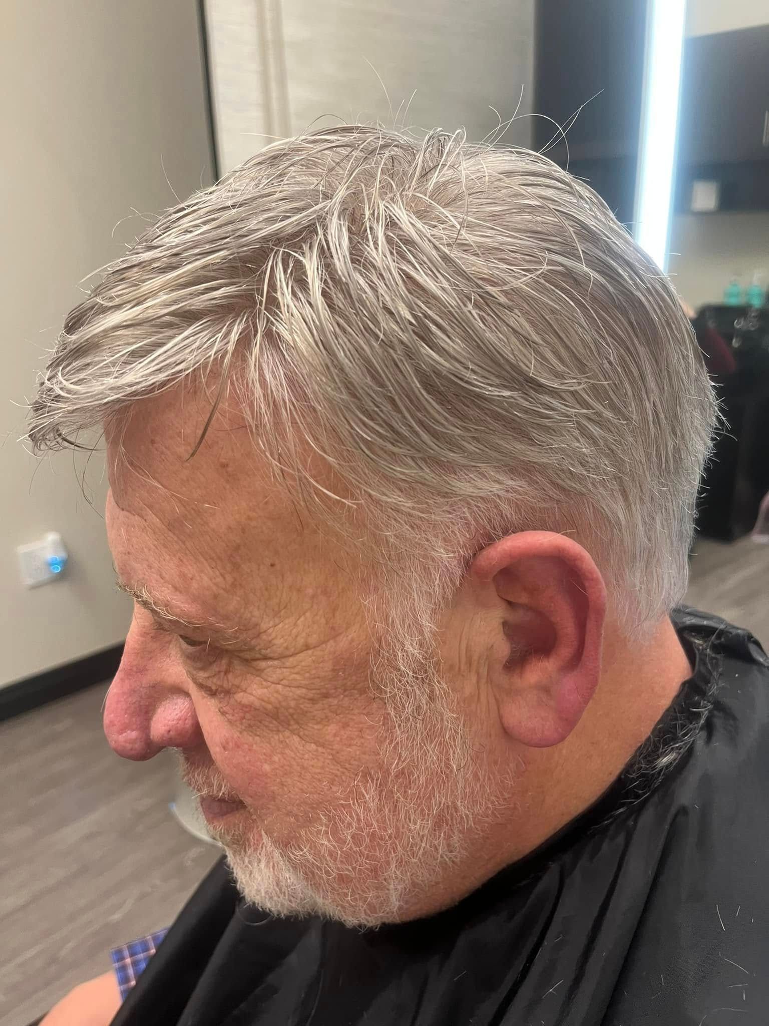 A person in a barber’s cape with hair and beard covered in small, white, loose hair clippings after a haircut.
